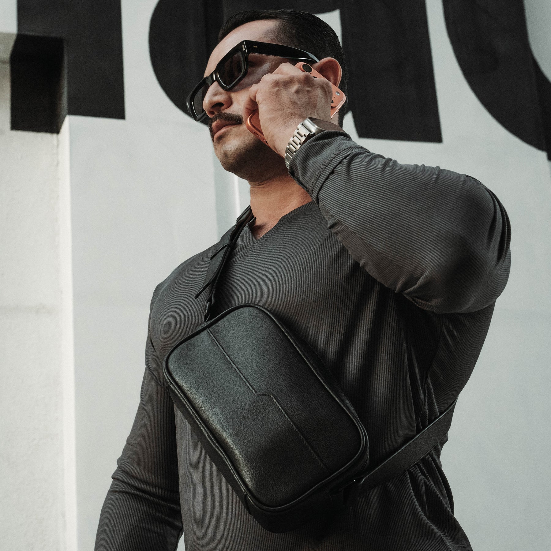 A man wearing the SANDMARC black leather crossbody sling across his chest, styled with a dark gray long-sleeve shirt, black sunglasses, and a Appl Watch Ultra against a white wall with black graphic lettering.