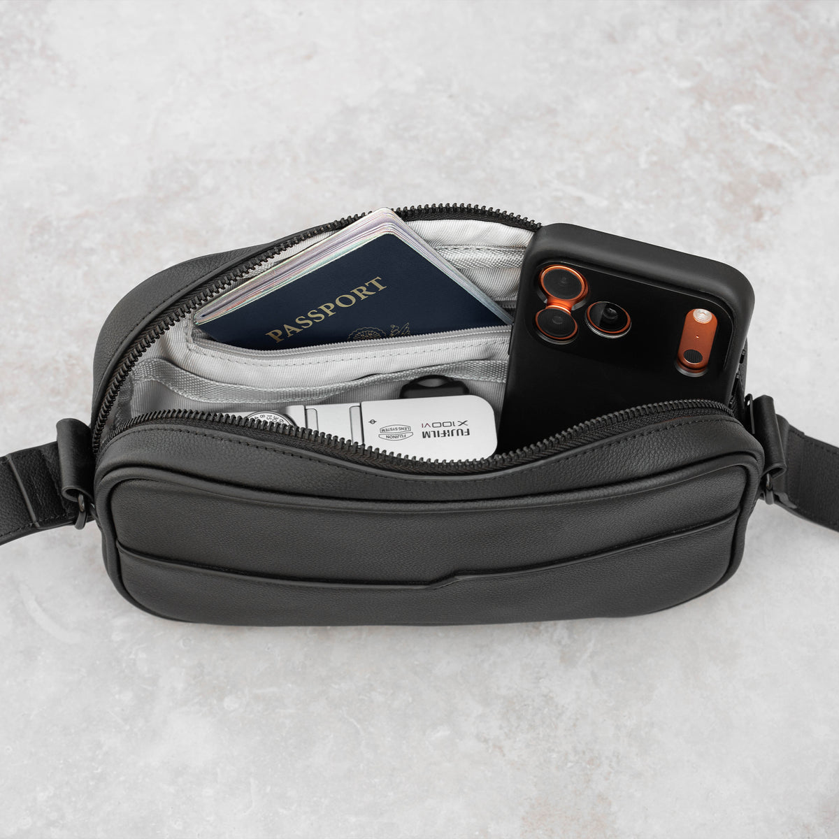 A black leather crossbody bag shown from above on a light gray textured surface, with its zipper open to display contents including a passport, Fujifilm camera, and an iPhone.