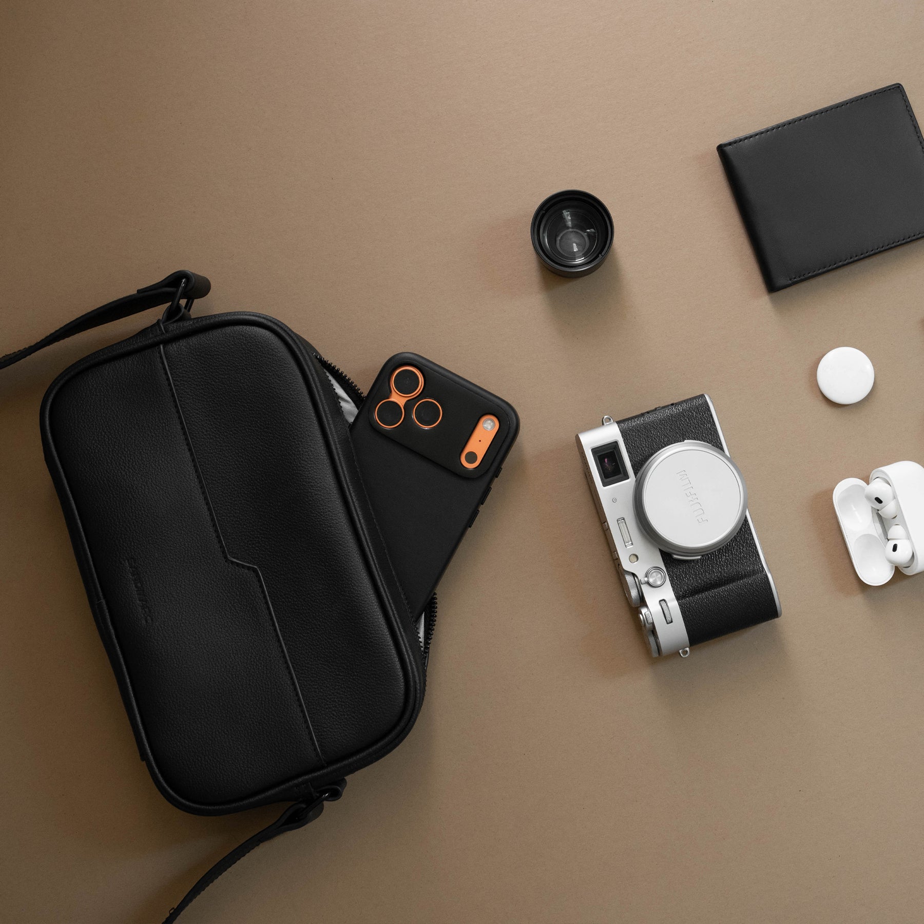A black zippered camera bag laid flat on a warm beige surface, styled with an iPhone, SANDMARC lens, wallet, AirTag, AirPods, and a compact camera.