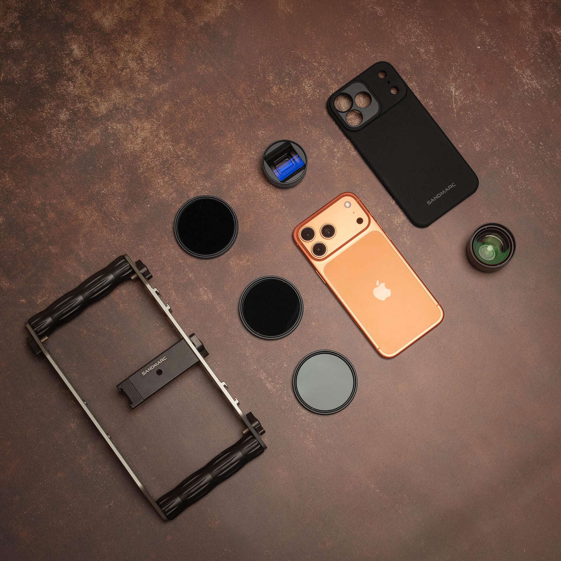 Overhead flat lay of the SANDMARC Film Edition on a dark rust-textured surface, showing the Film Rig, Anamorphic Lens, three ND Hybrid Filters, Telephoto 2x Lens, SANDMARC Standard Case, and iPhone 17 Pro laid out