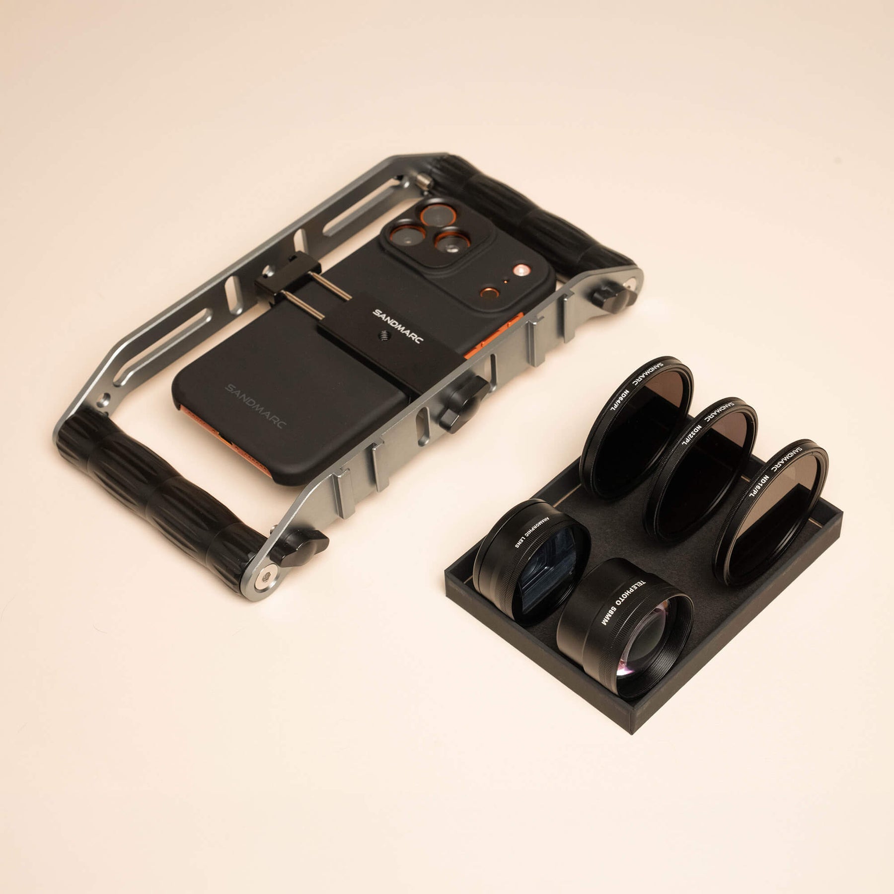 SANDMARC Film Rig holding an iPhone in a SANDMARC Standard Case on a cream background, positioned beside a black foam tray holding the Anamorphic Lens, Telephoto 58mm Lens, and three SANDMARC ND Hybrid Filters standing upright