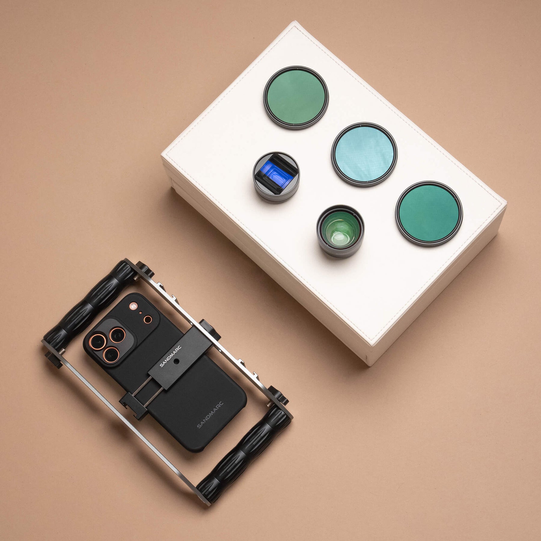 Overhead flat lay on a warm tan surface showing the SANDMARC Film Rig holding an iPhone in the SANDMARC Standard Case, beside a white box displaying the Anamorphic Lens, Telephoto 2x Lens, and three SANDMARC Hybrid Filters arranged on the lid
