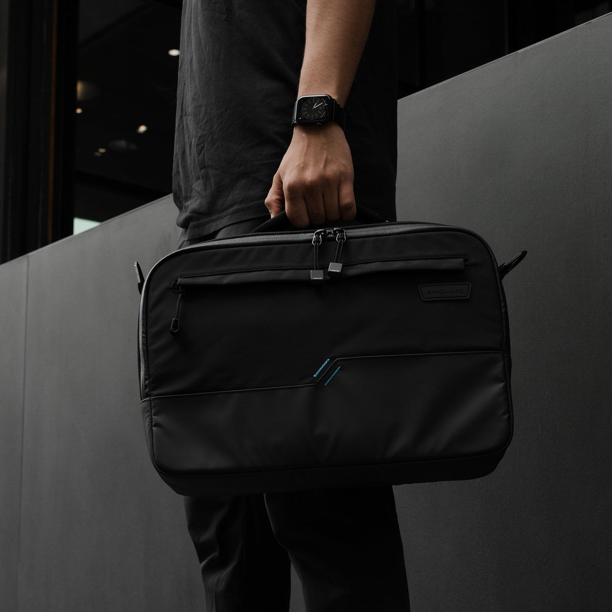 A person in a black shirt and pants holds a black SANDMARC messenger bag with a small blue accent stripe on the front, set against a dark gray modern wall.