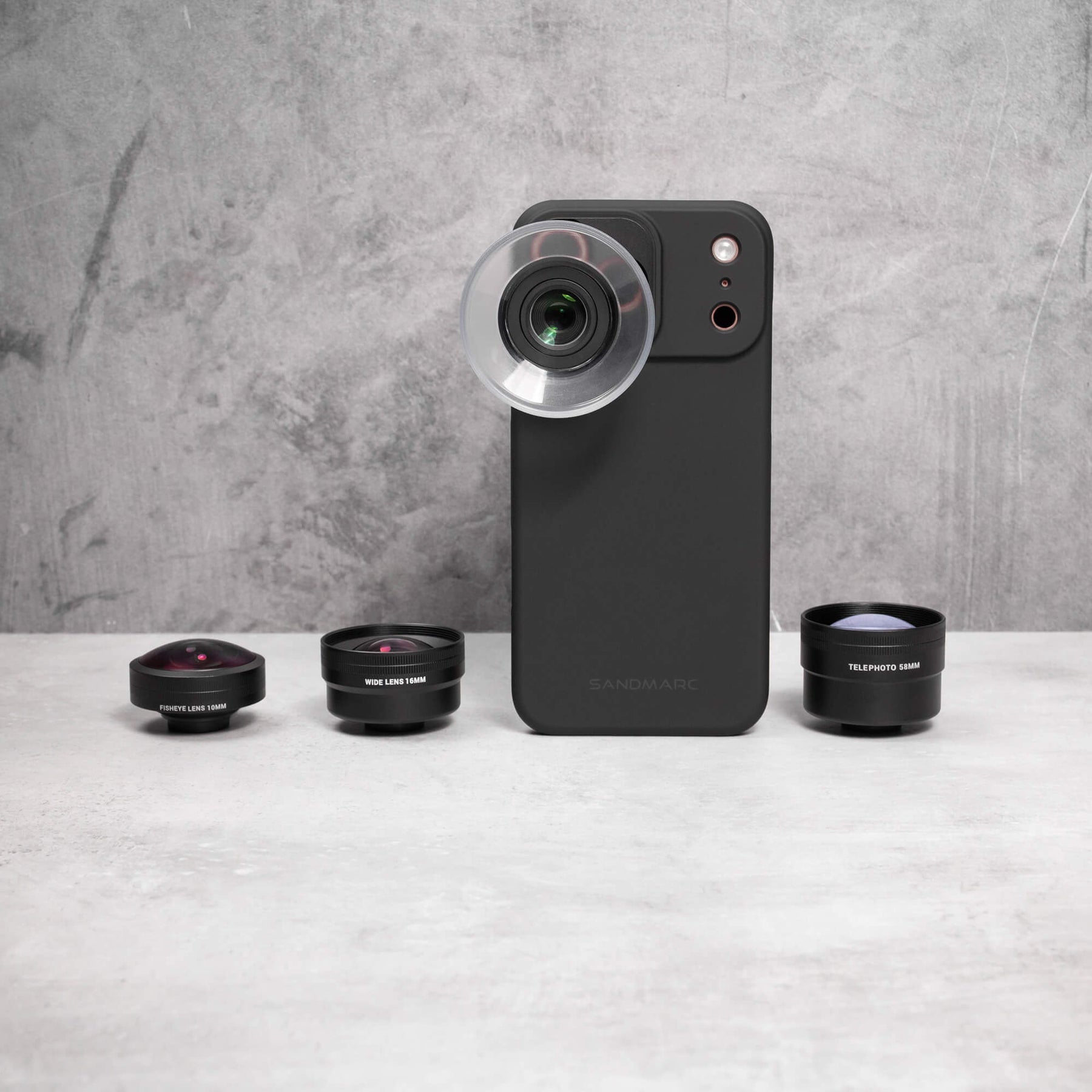 SANDMARC Standard Case standing upright with a macro lens attached, surrounded by three additional lenses — Fisheye, Wide, and Telephoto 58mm — laid out on a concrete surface