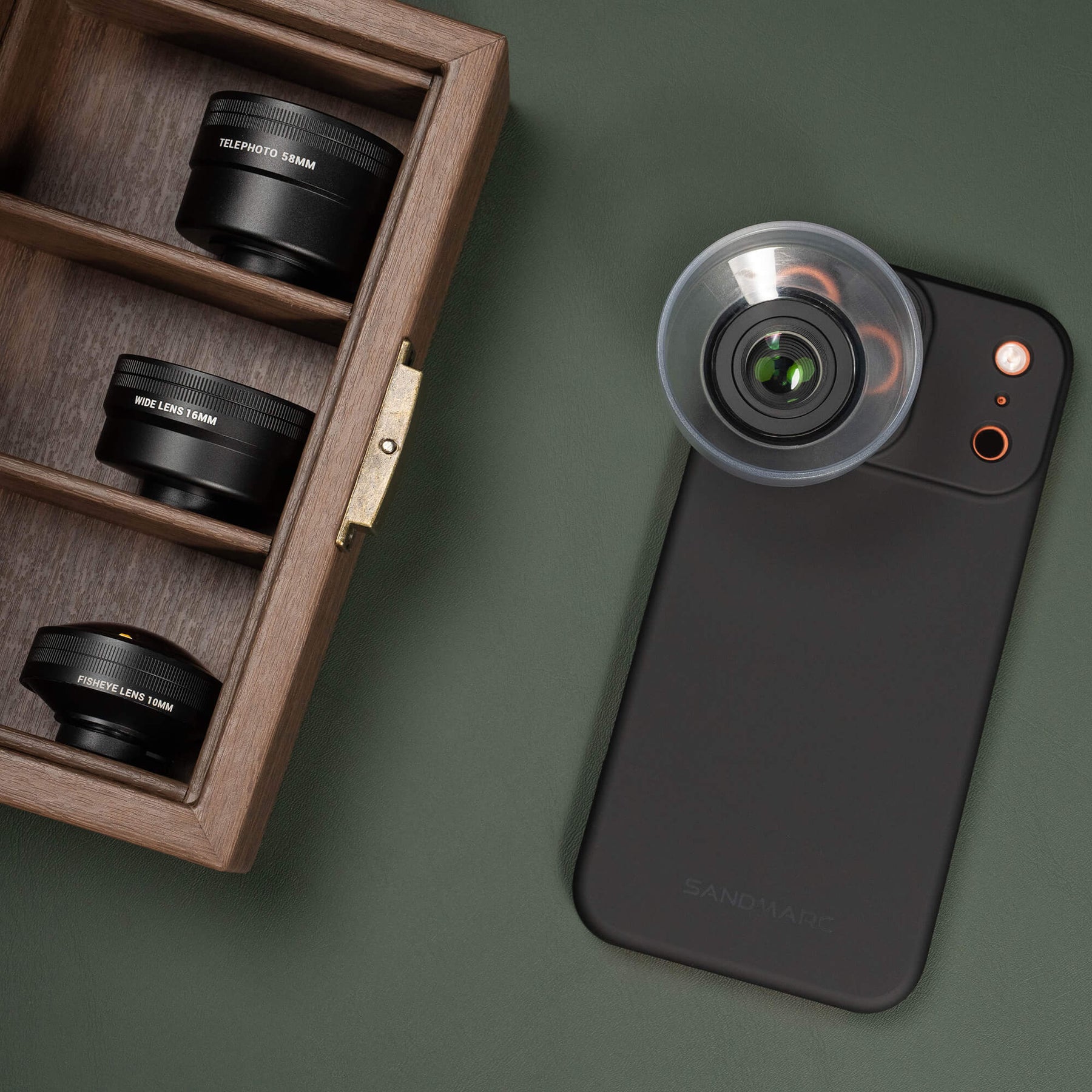 SANDMARC Standard Case with a macro 25mm lens attached lying on a dark green surface, beside an open wooden box containing three lenses — Telephoto 58mm, Wide, and Fisheye Lens