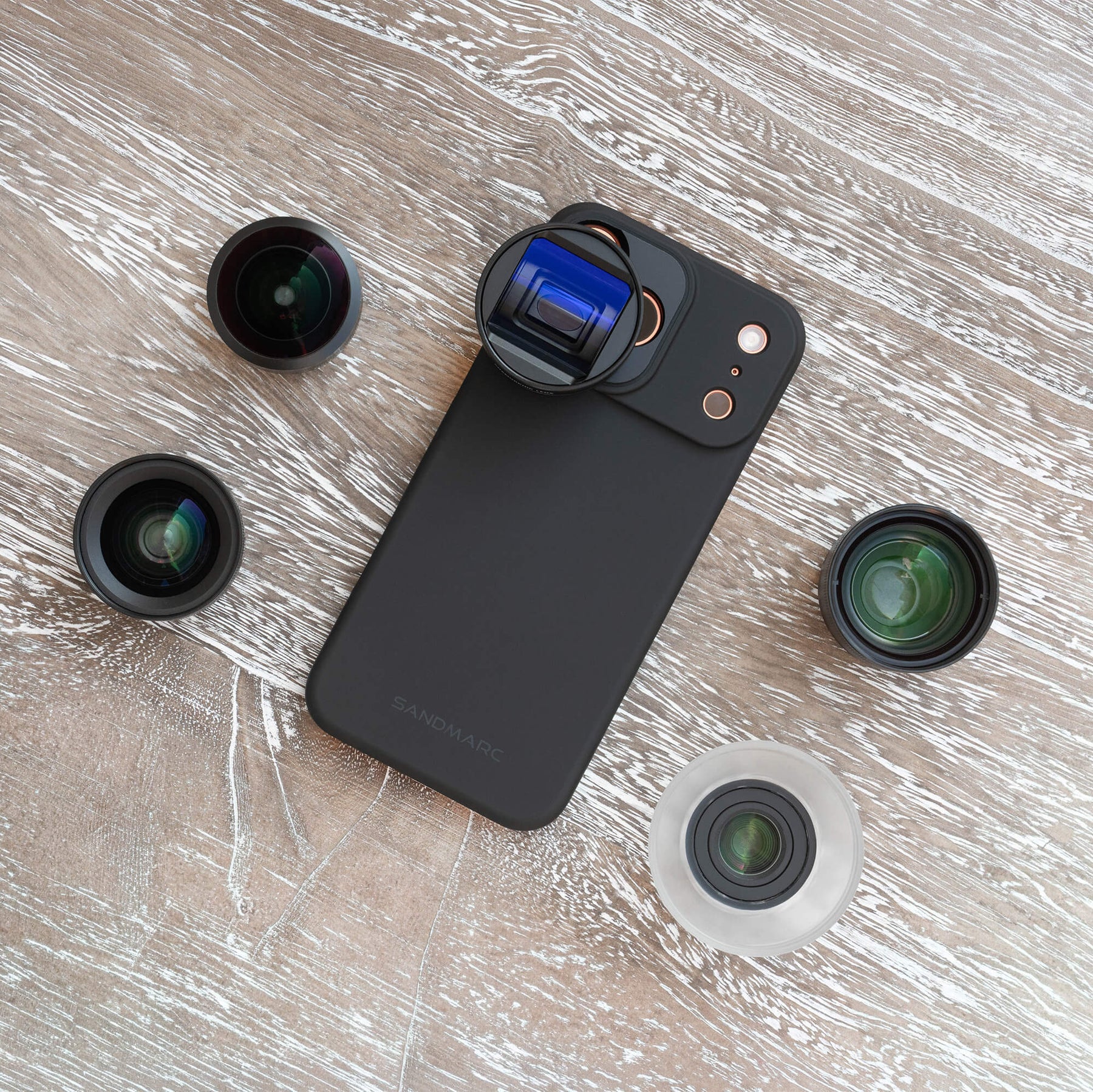 Overhead flat lay of the SANDMARC Pro Plus Edition on a whitewashed wood surface, showing a SANDMARC black Standard Case with Anamorphic Lens attached, surrounded by four additional lenses — Fisheye, Wide, Telephoto, and Macro