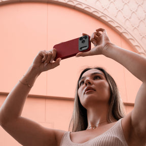 Woman holding an iPhone in the SANDMARC Pro Leather Case in red against their black leather jacket, showing the full-grain leather texture and aluminum camera mount