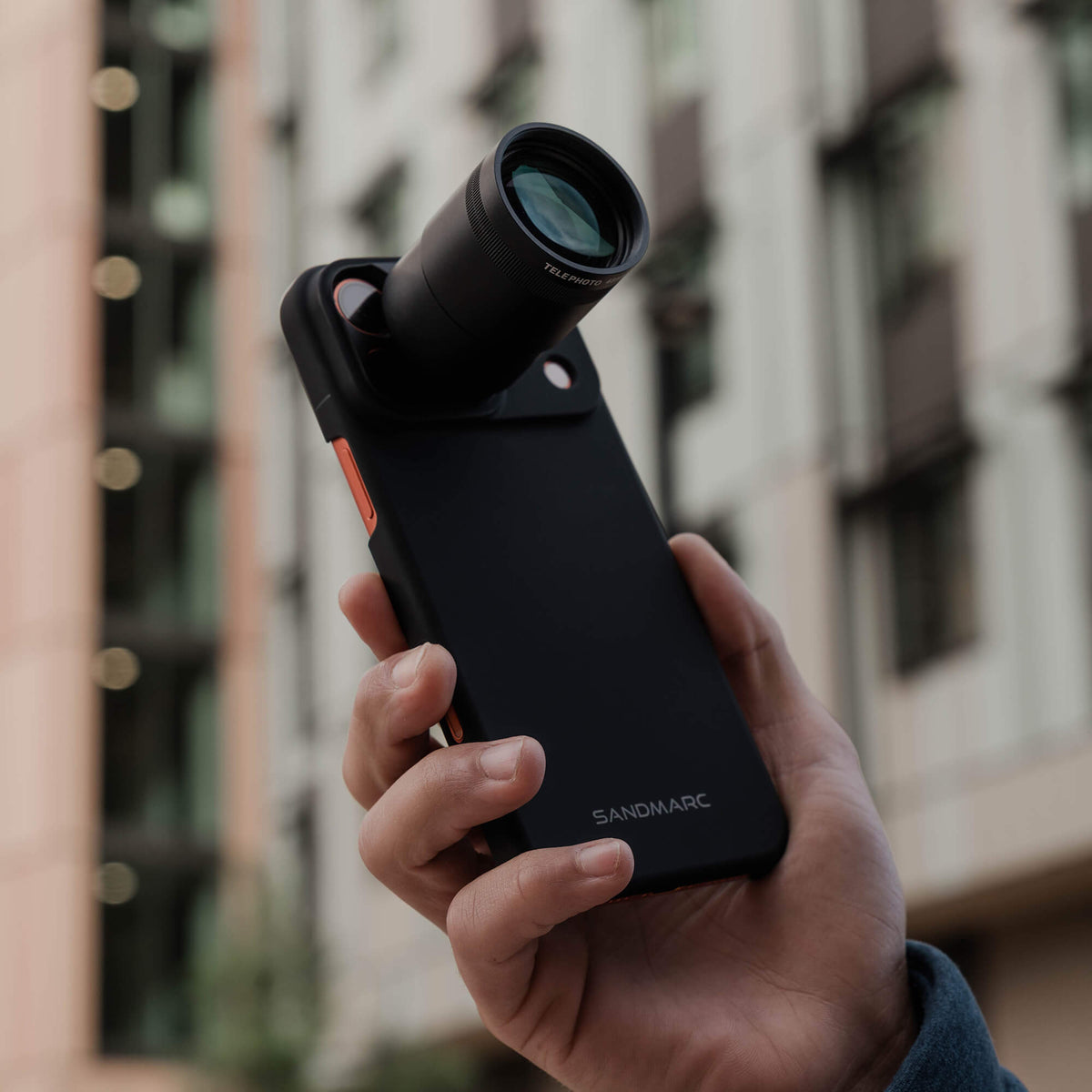 Hand holding an iPhone with a mounted 72mm SANDMARCED lens against an urban background