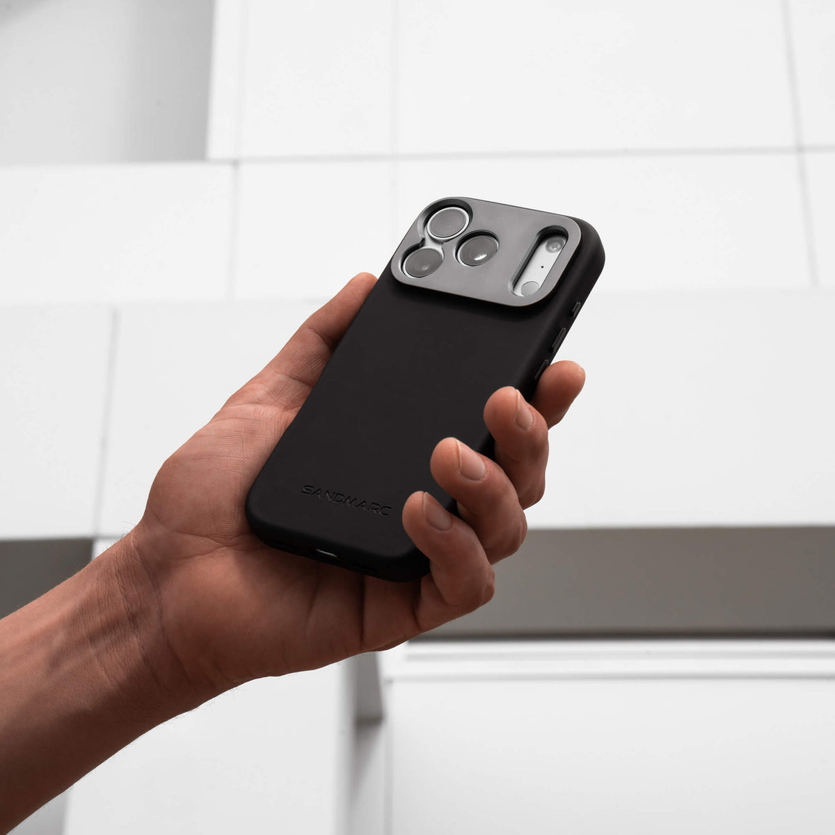 Hand holding an iPhone in the SANDMARC Pro Case in black against a white tiled background, showing the aluminum camera mount and triple camera cutout with natural titanium finish