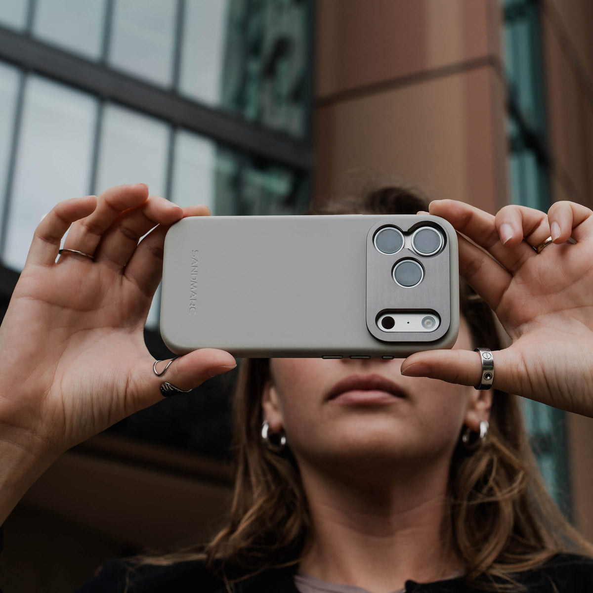 Woman holding an iPhone in the SANDMARC Pro Case in stone horizontally up to her face outdoors in front of a modern building, wearing silver rings and hoop earrings, showing the SANDMARC logo and camera mount