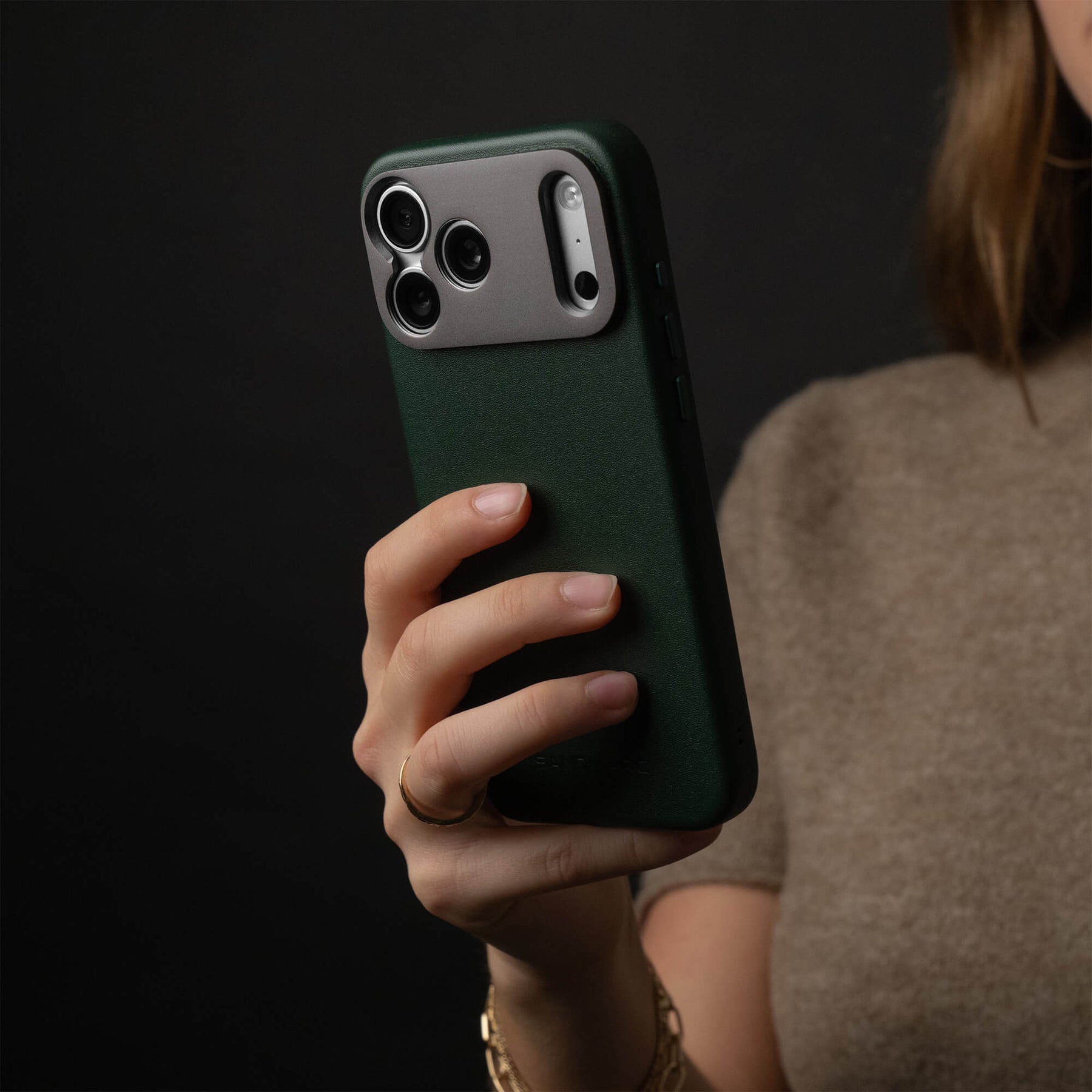 Woman holding an iPhone in the SANDMARC Pro Leather Case in forest green against a dark background, wearing a gold ring and bracelet with a taupe knit top, showing the full-grain leather texture