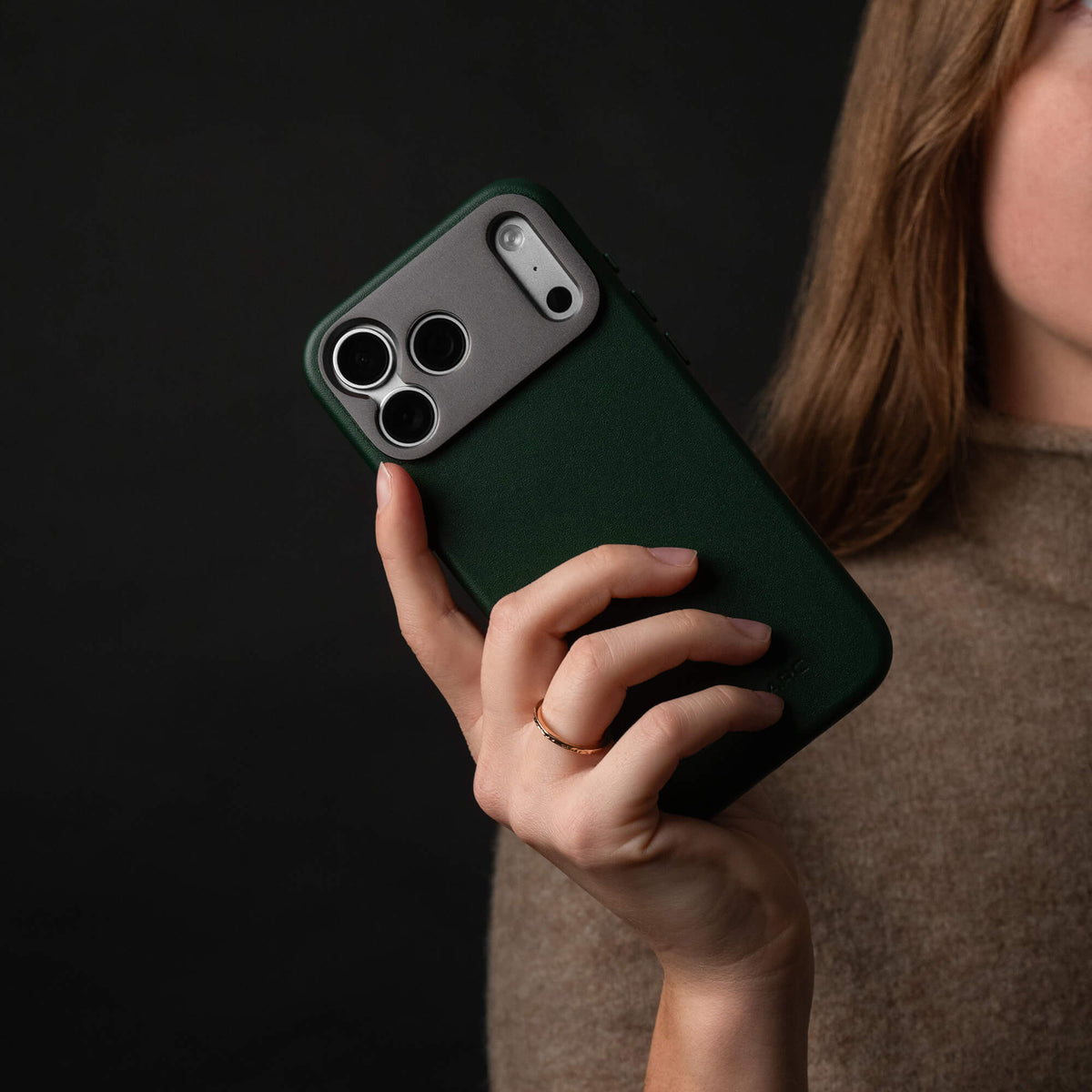 Woman holding an iPhone in the SANDMARC Pro Leather Case in forest green close to her face against a dark background, wearing a gold ring and taupe knit top, showing the aluminum camera mount and triple lens cutout