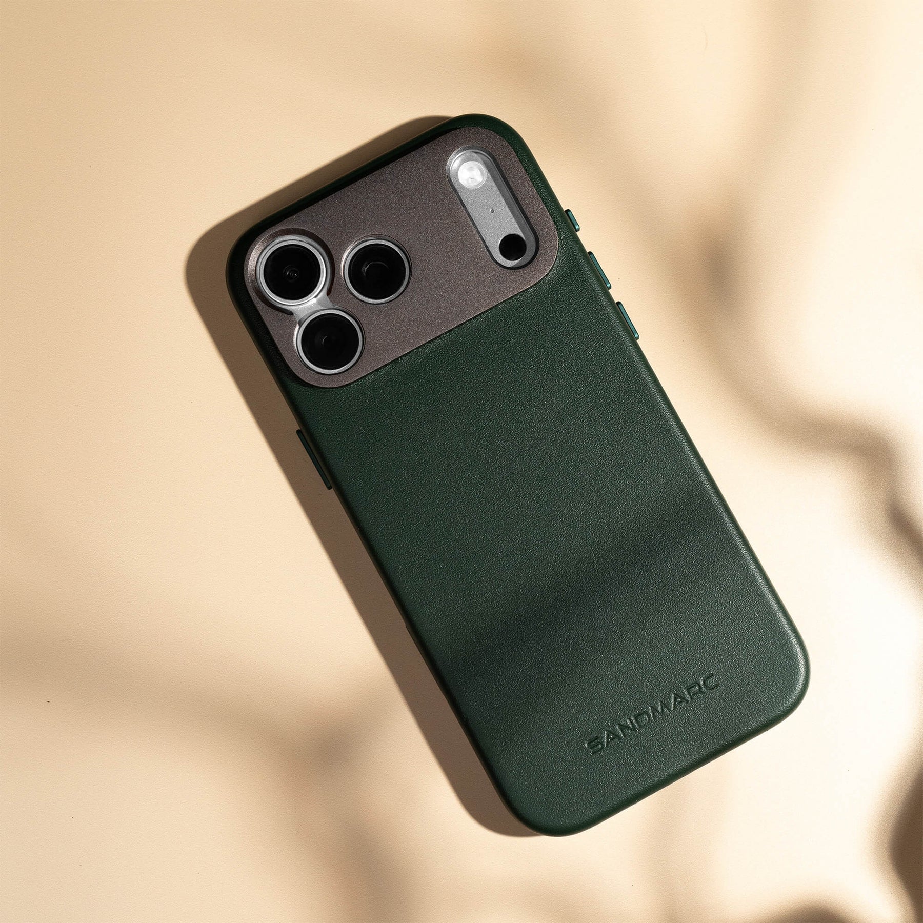 SANDMARC Pro Leather Case in forest green on iPhone lying on a warm cream surface with natural leaf shadows, showing the SANDMARC logo embossed on the leather