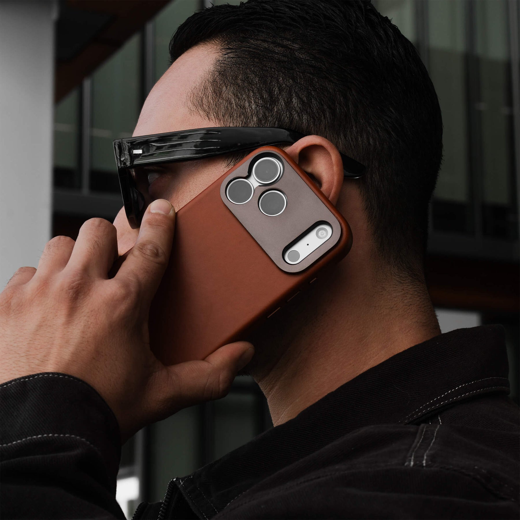 Man holding an iPhone in the SANDMARC Pro Leather Case in tan up to his ear against a modern building exterior, wearing black sunglasses and a black jacket, showing the aluminum camera mount and triple lens cutout