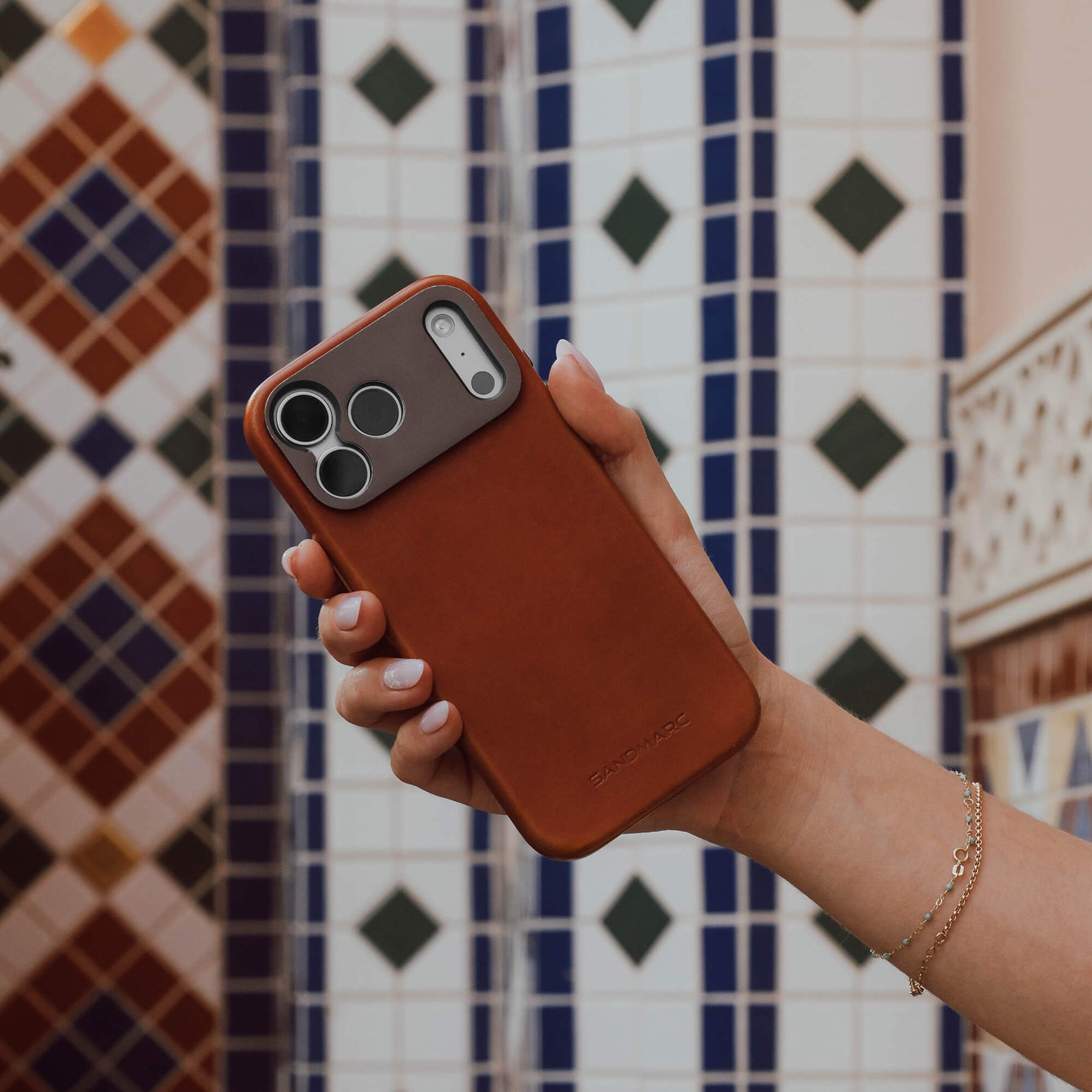 Woman's hand holding an iPhone in the SANDMARC Pro Leather Case in tan against a colorful mosaic tile wall, wearing a delicate gold chain bracelet and showing the SANDMARC logo embossed on the leather