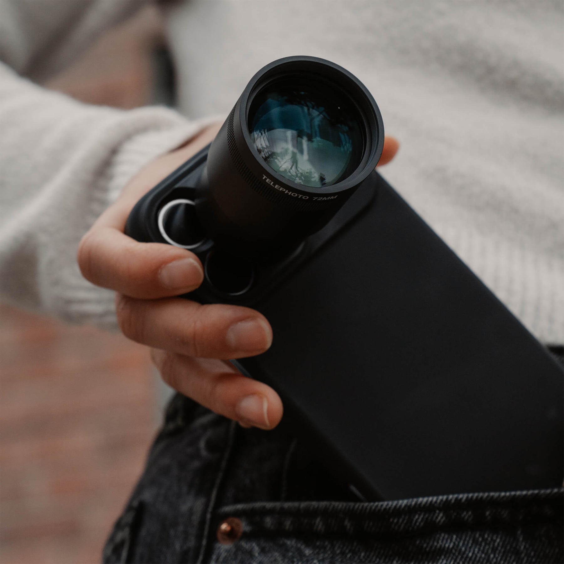 Hand gripping an iPhone 18 Pro Max in a SANDMARC Black Standard Case with the Telephoto Tetraprism 72mm Lens pointed toward camera, shown being pulled from a denim jeans pocket outdoors, wearing a grey knit sweater