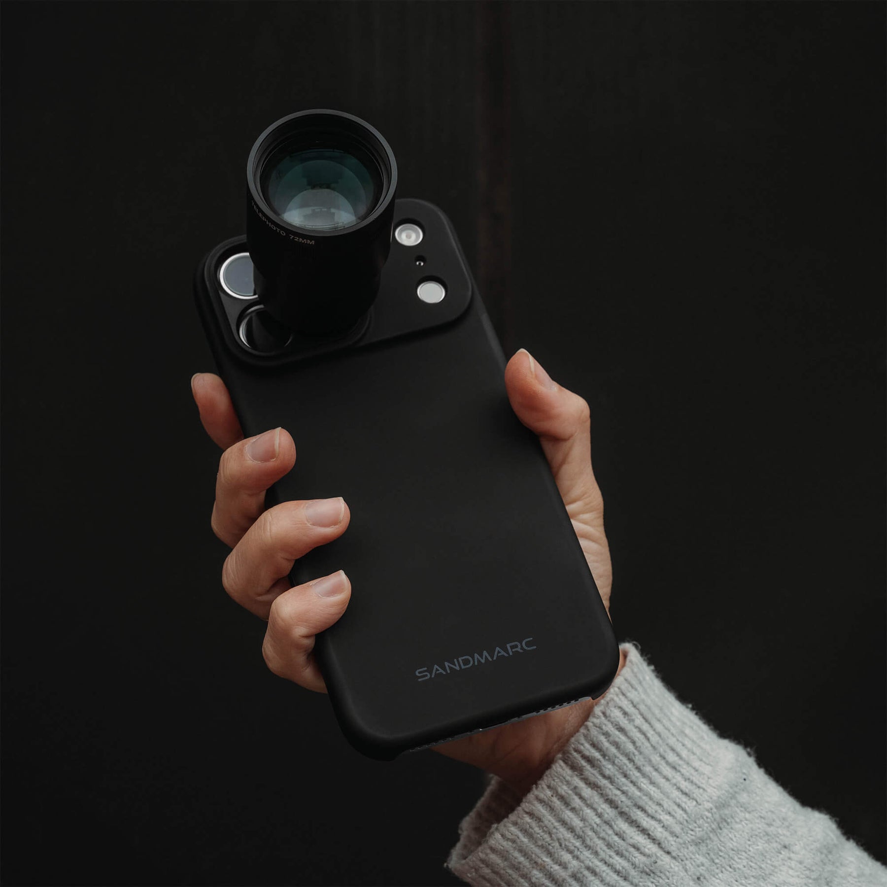 Hand holding an iPhone 18 Pro Max in a SANDMARC Black Standard Case with the Telephoto Tetraprism 72mm Lens attached, held up against a pure black background, wearing a white knit sweater and showing the SANDMARC logo on the case