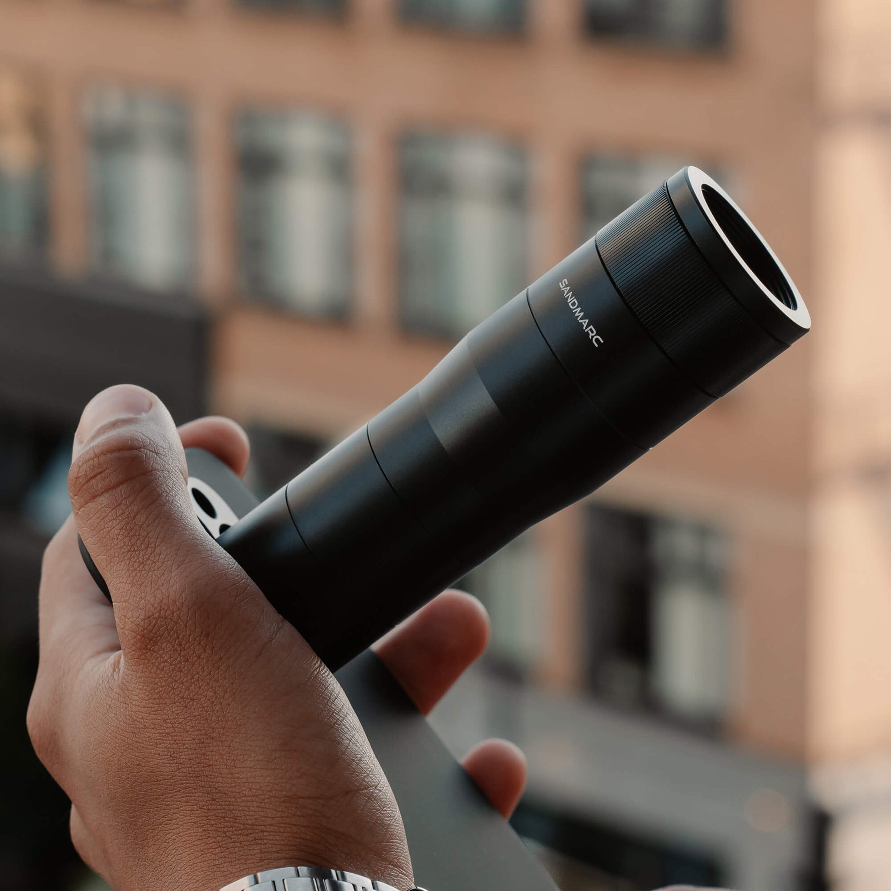 SANDMARC Telephoto 6x Lens, held up by a hand against a blurred building facade background
