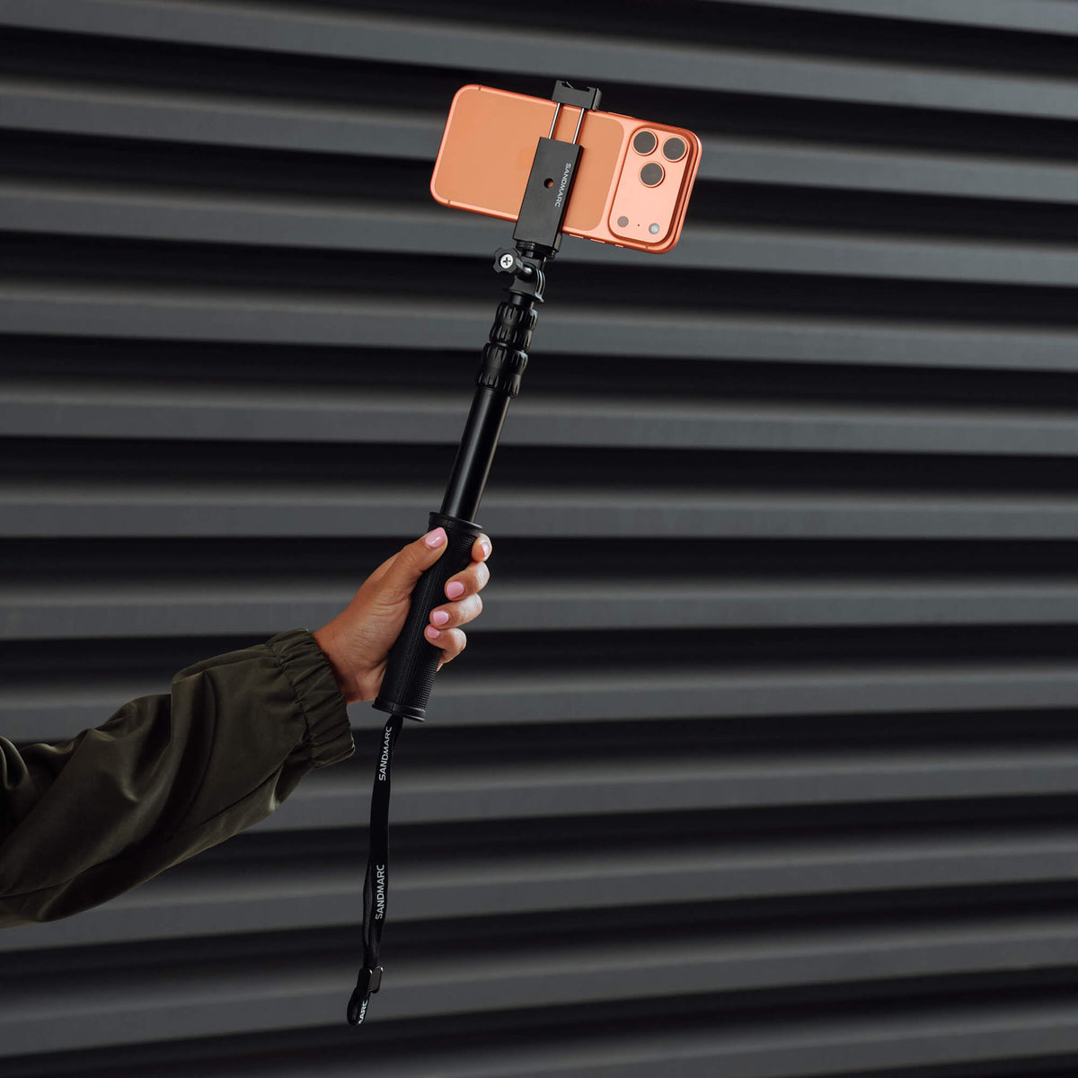 Photographer holding SANDMARC Pole with iPhone 17 Pro Max attached on dark striped background