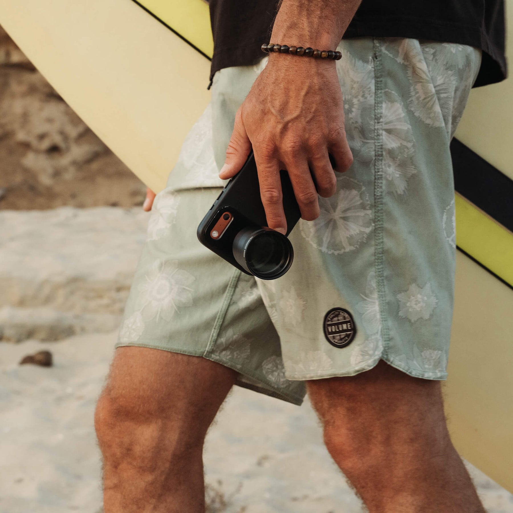 Close-up of a man's hand carrying an iPhone in a black SANDMARC Pro Leather case with a Telephoto 96mm lens attached at his side, wearing board shorts and a beaded bracelet at the beach