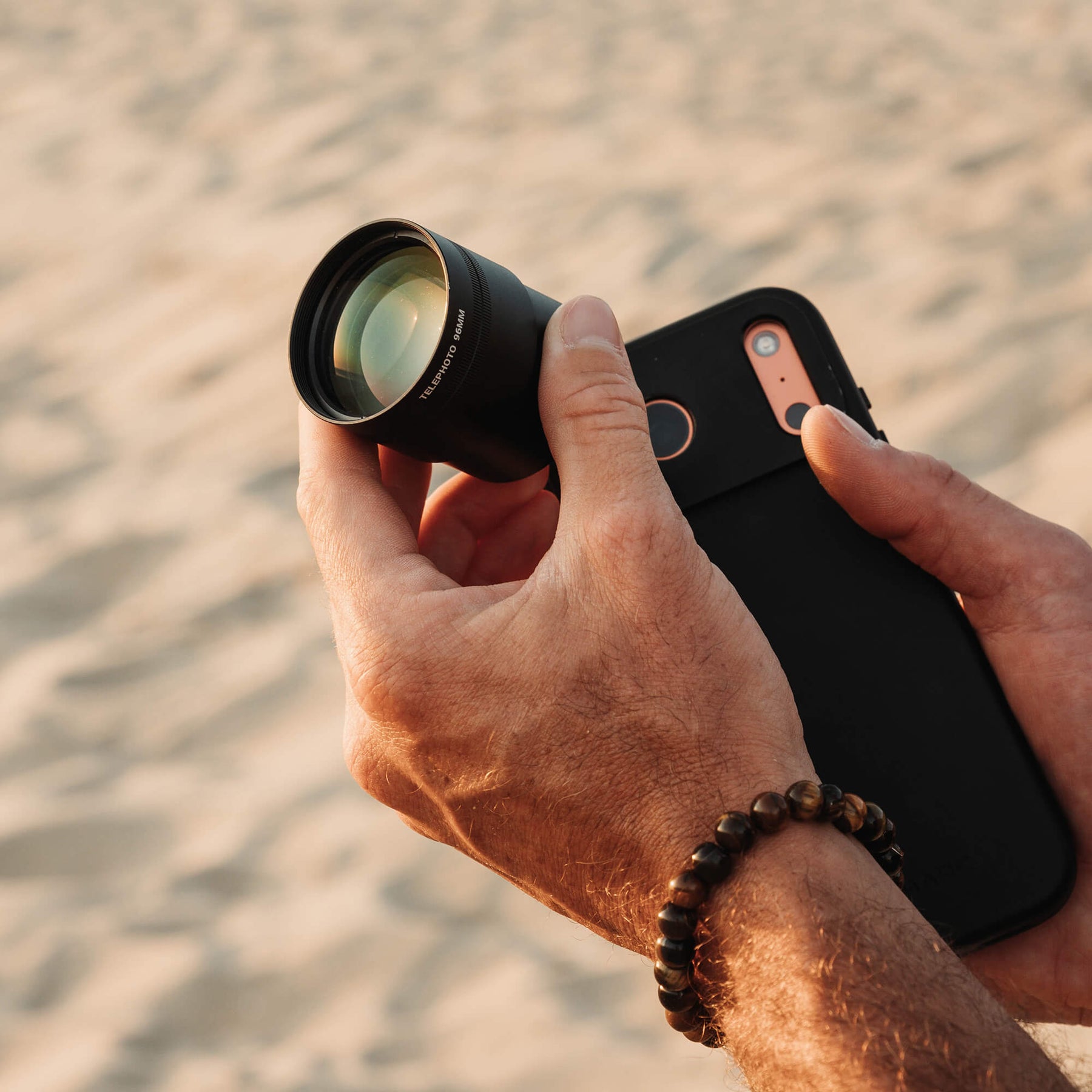 Close-up of a man's hand holding an iPhone in a black SANDMARC Pro Leather case with a Telephoto 96mm lens attached, photographed on a sandy beach at golden hour