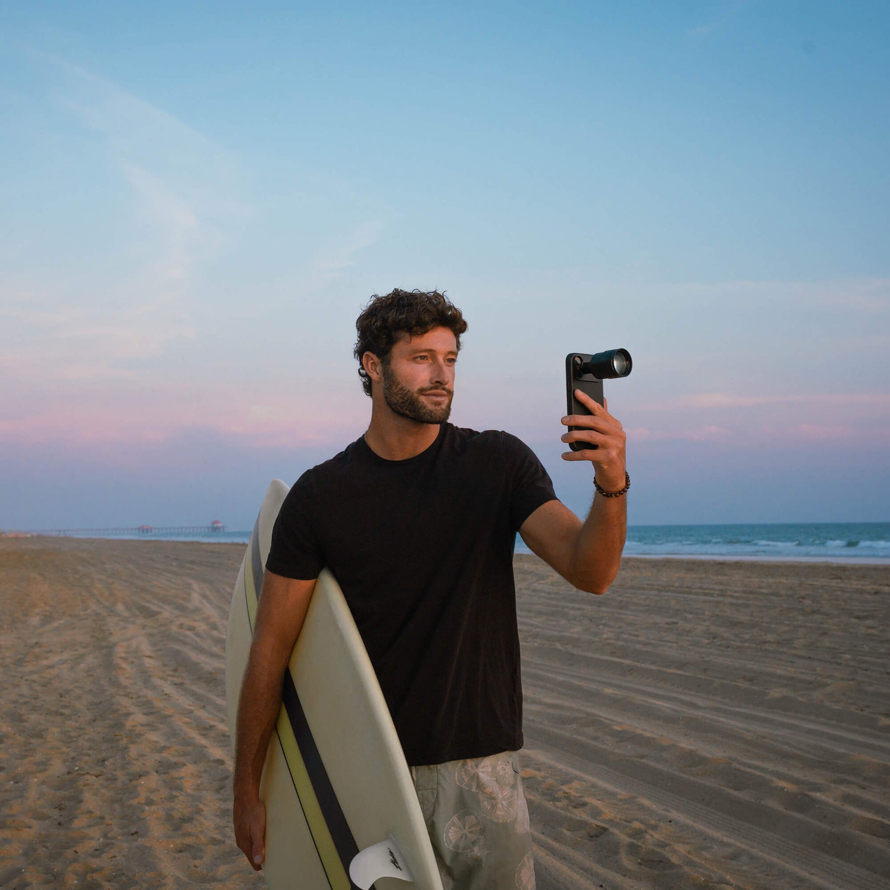 Man carrying a surfboard on a beach at sunset holding up an iPhone in a black SANDMARC Pro Leather case with a Telephoto 96mm lens attached, pastel pink and blue sky in the background