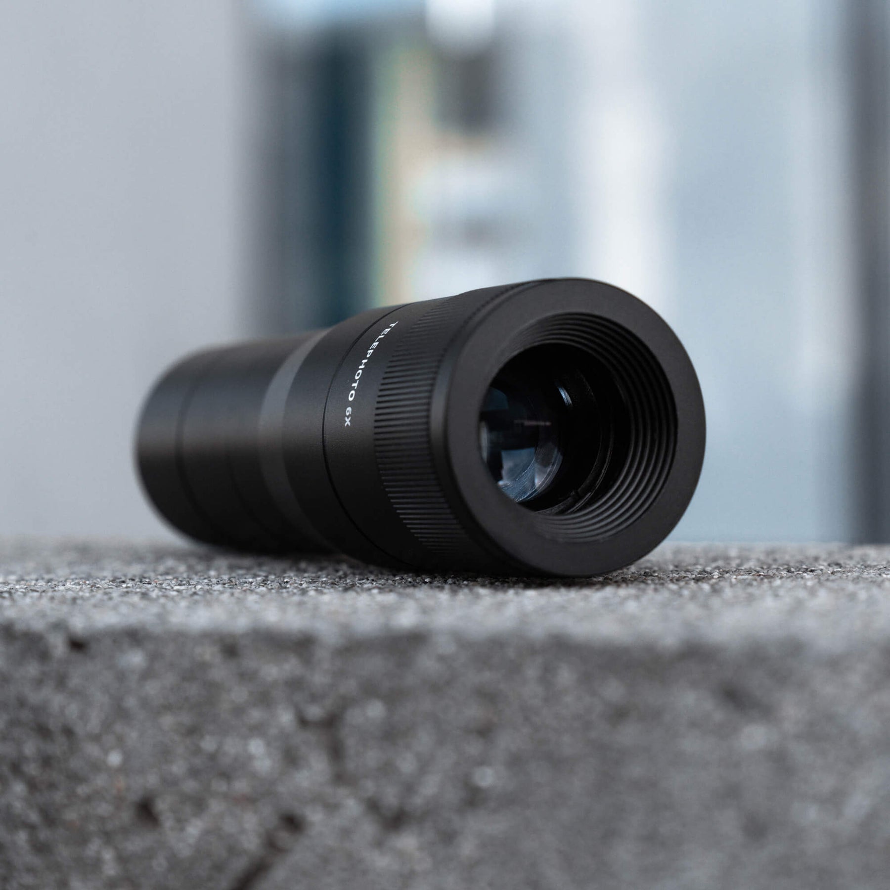 SANDMARC Telephoto 6x Lens, lying on its side on a concrete ledge, showing the front glass element and blurred building background