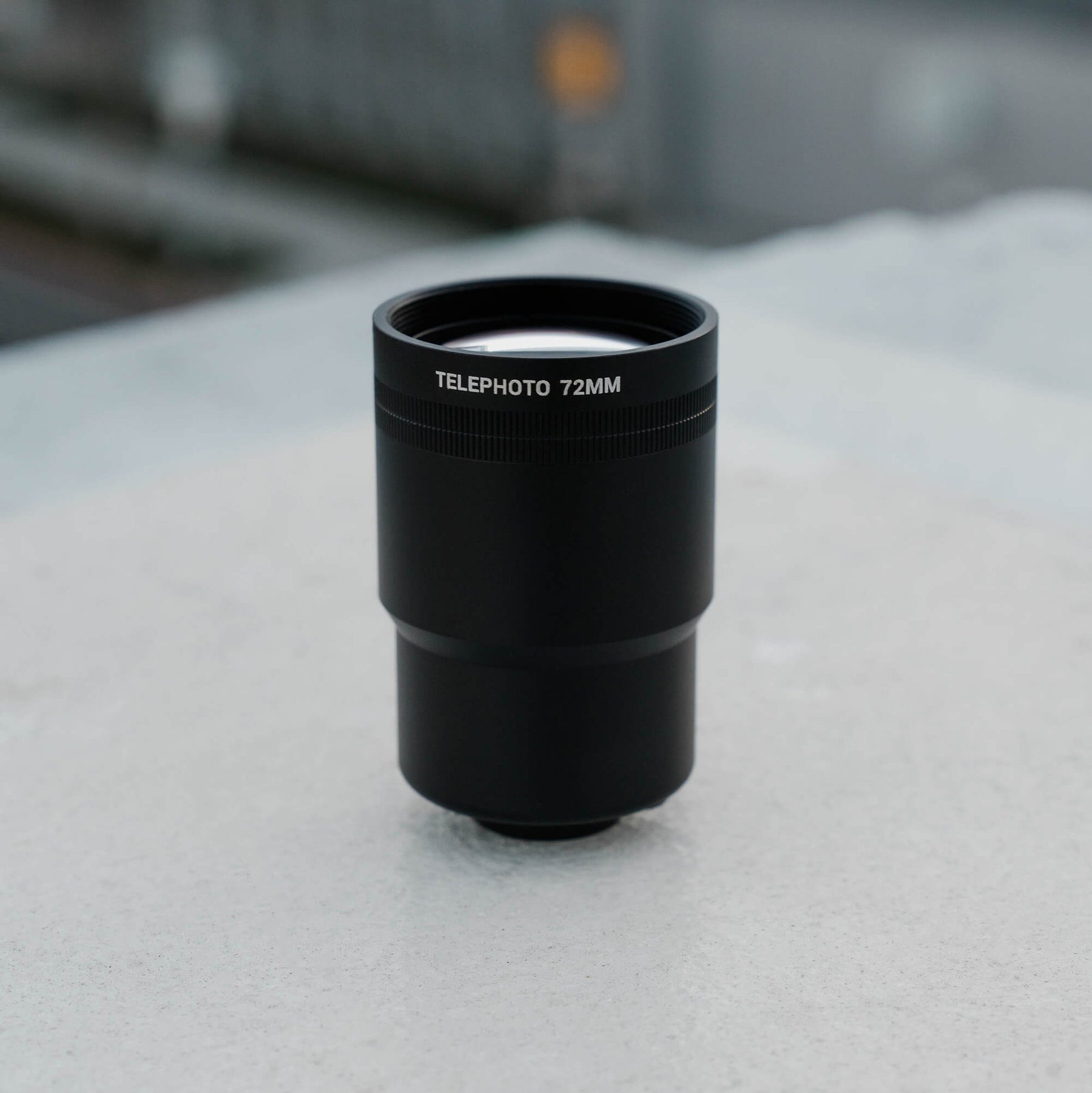 SANDMARC Tetraprism lens with '72MM' marking on a light gray concrete.
