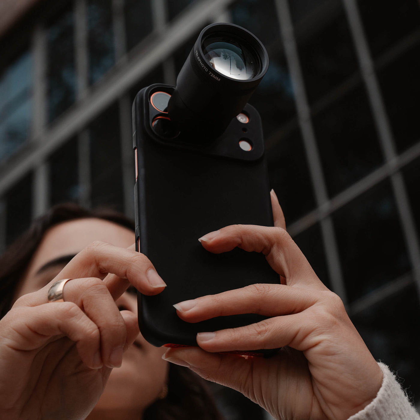 Person holding a iPhone with the 72mm camera lens attachment against a blurred cityscape background.