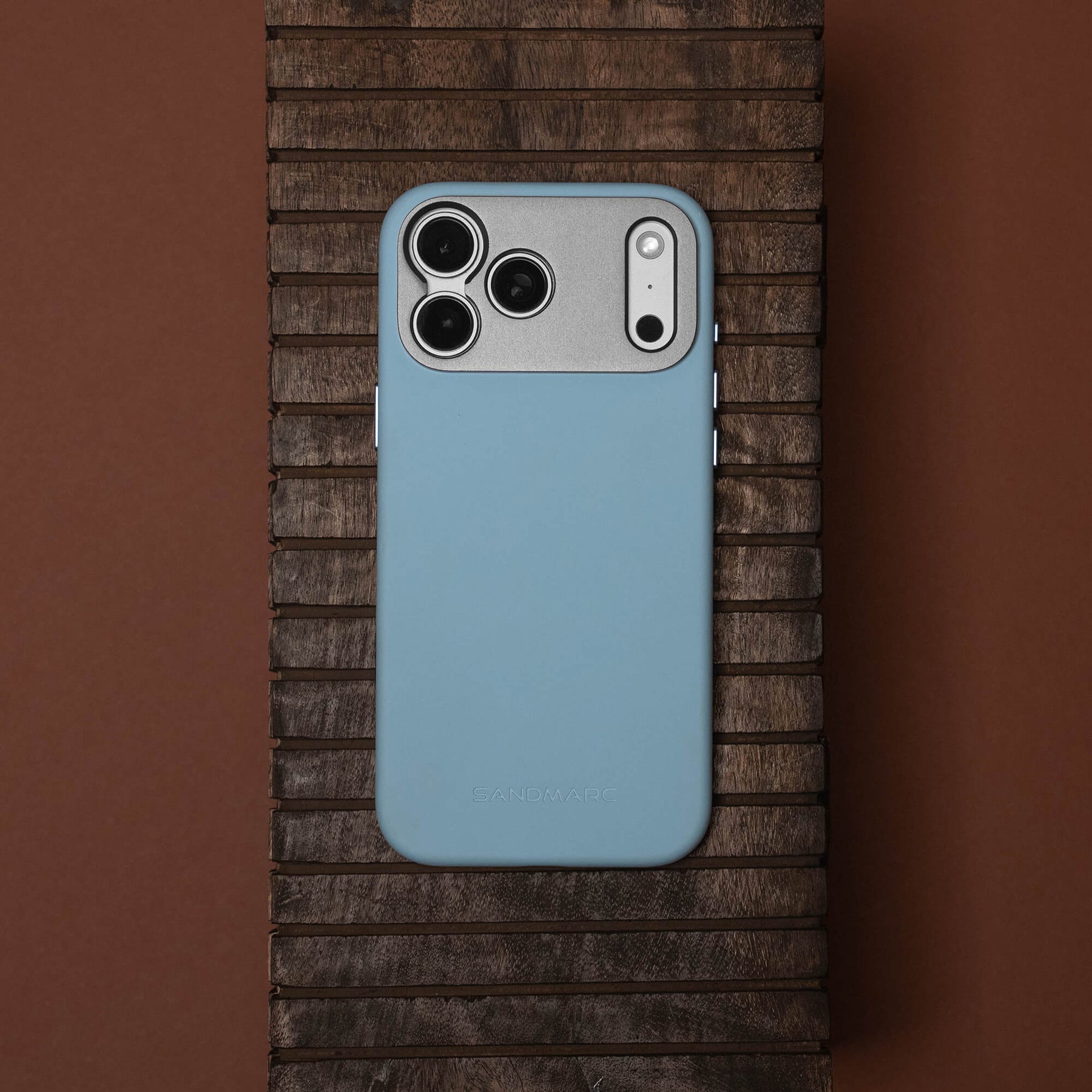 SANDMARC Pro Case for iPhone 18 Pro in Glacier blue, lying flat on a dark wood slatted surface with a brown background.