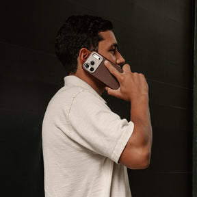 Woman holding an iPhone in the SANDMARC Pro Leather Case in brown against a dark background, wearing a gold ring and a taupe knit top, showing the leather texture and camera mount