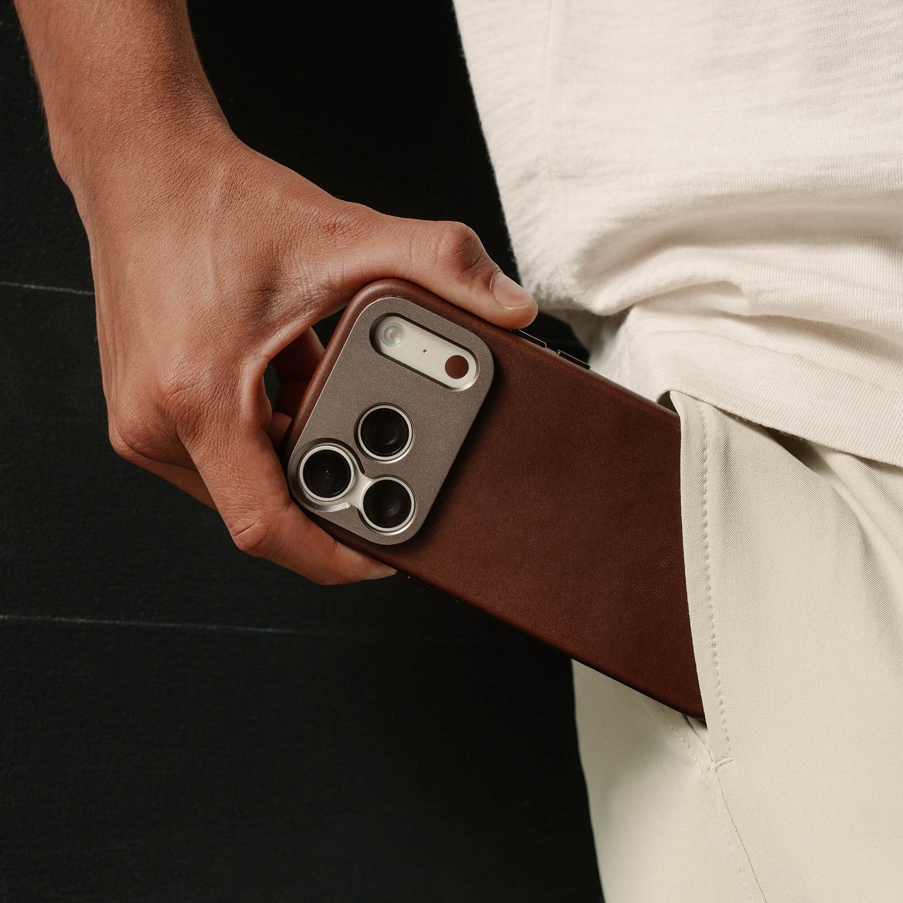 Close-up of a hand pulling an iPhone in the SANDMARC Pro Leather Case in brown out of a pocket, showing the aluminum camera mount and triple lens cutout