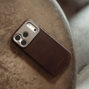 Woman holding an iPhone in the SANDMARC Pro Leather Case in brown against a dark background, wearing a gold ring and a taupe knit top, showing the leather texture and camera mount