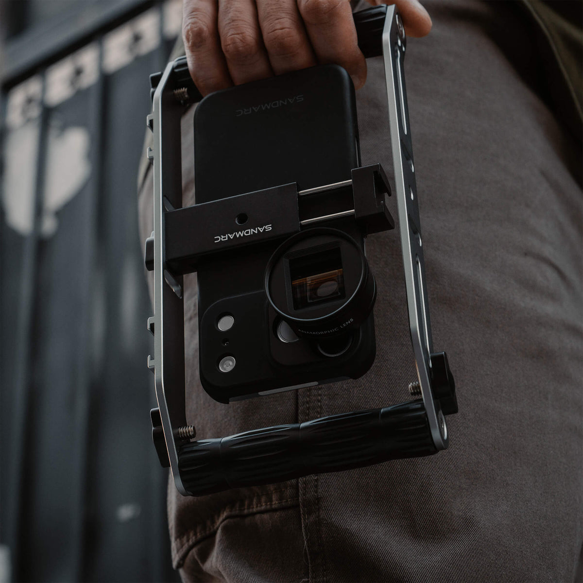 Man carrying the SANDMARC Film Rig with an iPhone 18 Pro Max mounted face-down and the Anamorphic 1.33x Lens attached, showing the SANDMARC logo on both the case and rig bracket, held at his side outdoors against a blue garage door #type_anamorphic 1.33x