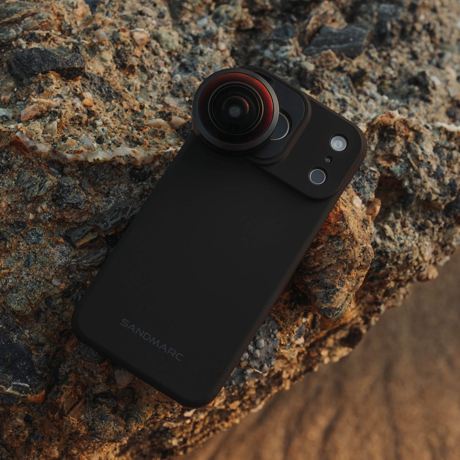 SANDMARC Fisheye Lens 10mm attached to an iPhone 18 Pro Max in a SANDMARC Black Standard Case resting on textured coastal rocks at golden hour, showing the red-tinted lens element and SANDMARC logo
