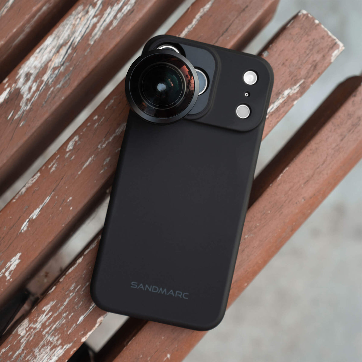 SANDMARC Fisheye Lens 10mm attached to an iPhone 18 Pro Max in a SANDMARC Black Standard Case lying on weathered red wooden slats outdoors, showing the SANDMARC logo and lens mounted to the camera