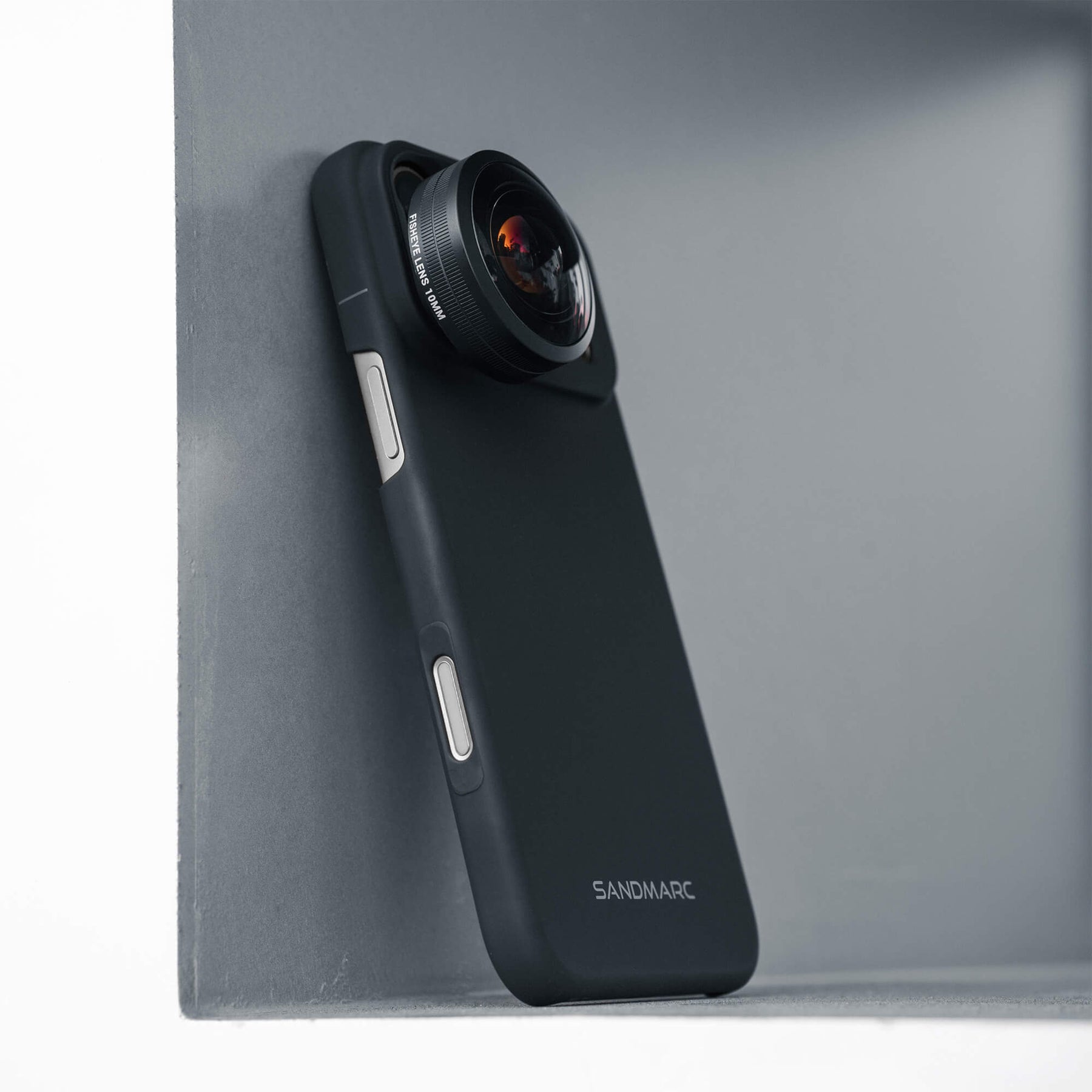 SANDMARC Fisheye Lens 10mm attached to an iPhone 18 Pro Max in a SANDMARC Black Standard Case leaning against a grey concrete corner, showing the wide curved glass element and lens barrel detail