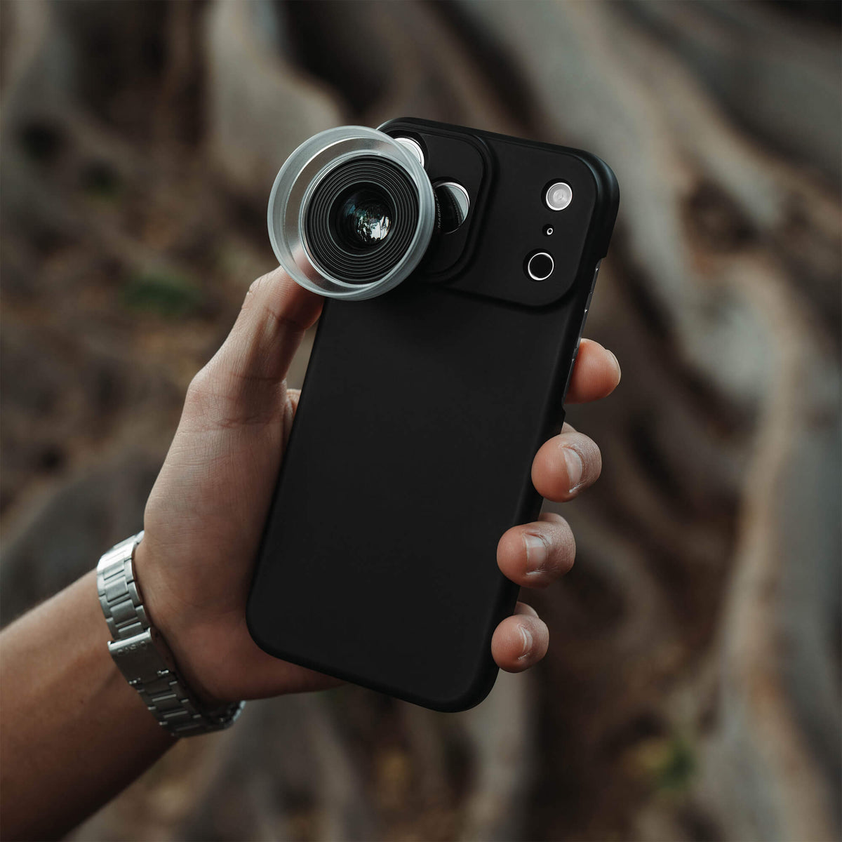 Man's hand holding an iPhone 18 Pro Max in a SANDMARC Black Standard Case with the Macro 25mm Lens attached, wearing a SANDMARC titanium watch band with blurred tree roots visible in the background #type_macro 25mm