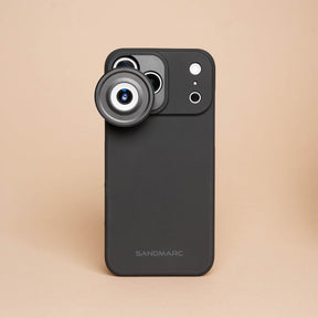 iPhone 18 Pro Max in a SANDMARC Black Standard Case with the Microscope Lens attached lying flat on a textured rust and grey concrete surface, showing the SANDMARC logo and circular lens element