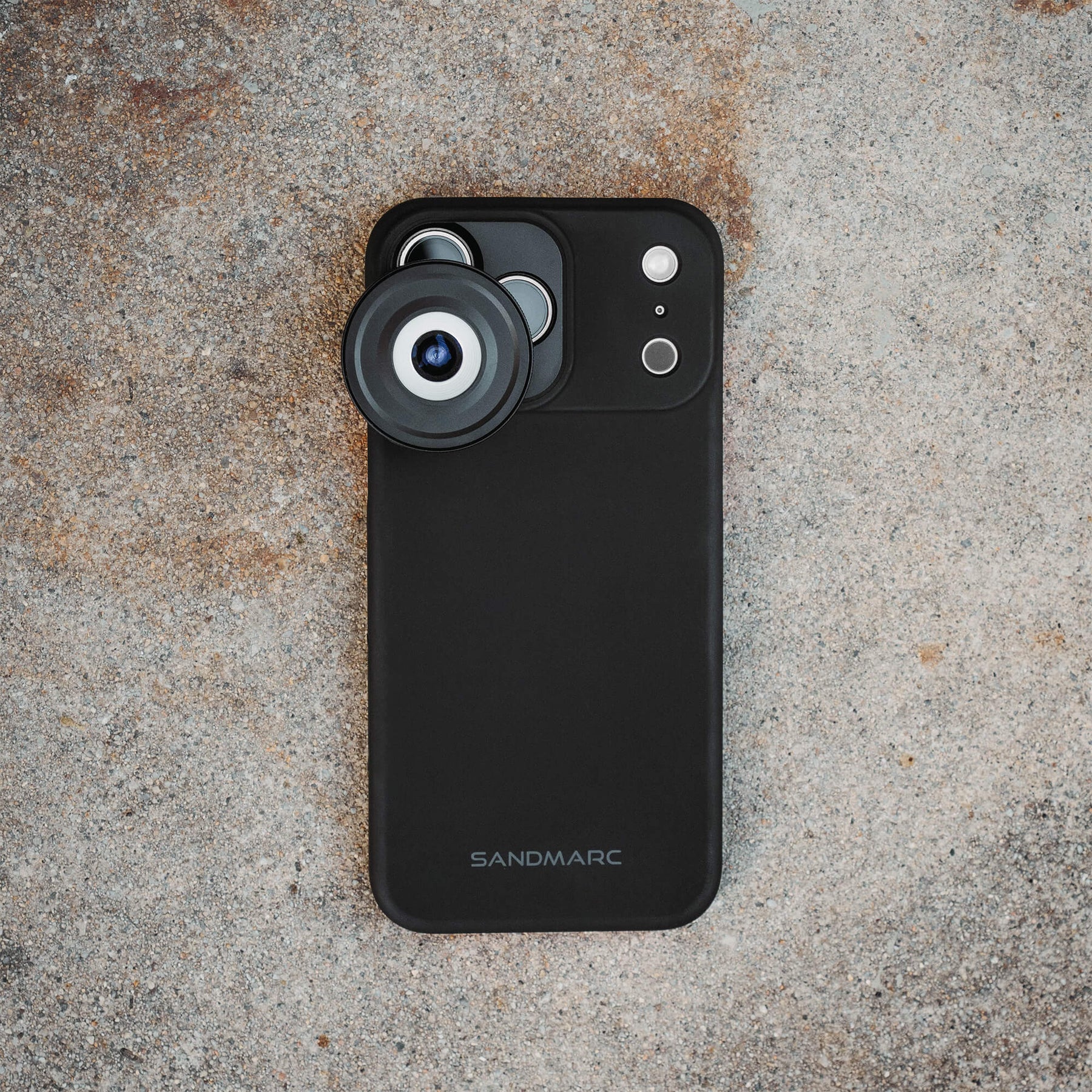 iPhone 18 Pro Max in a SANDMARC Black Standard Case with the Microscope Lens attached lying flat on a textured rust and grey concrete surface, showing the SANDMARC logo and circular lens element