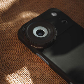 iPhone 18 Pro Max in a SANDMARC Black Standard Case with the Microscope Lens attached lying flat on a textured rust and grey concrete surface, showing the SANDMARC logo and circular lens element