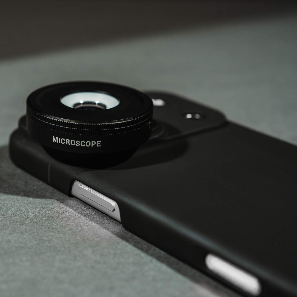 Close-up of the SANDMARC Microscope Lens attached to an iPhone 18 Pro Max in a SANDMARC Black Standard Case lying on a grey surface, showing the lens barrel engraving, knurled focus ring, and matte black finish
