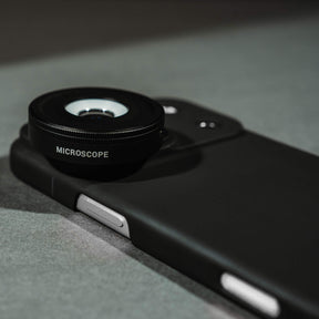 iPhone 18 Pro Max in a SANDMARC Black Standard Case with the Microscope Lens attached lying flat on a textured rust and grey concrete surface, showing the SANDMARC logo and circular lens element