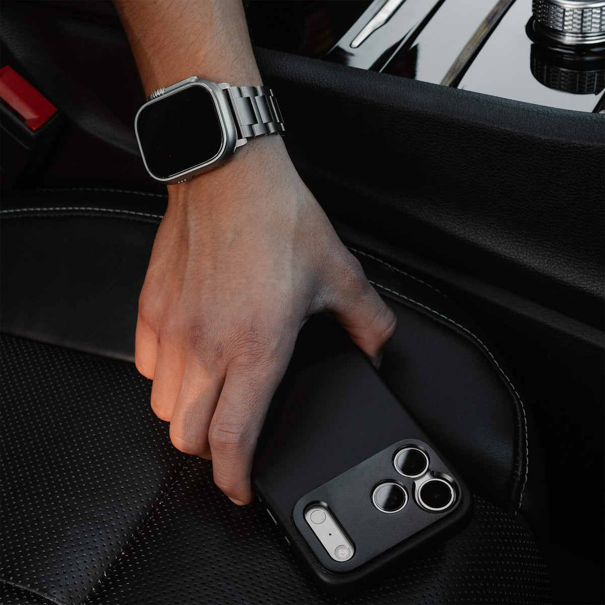 Man's hand picking up an iPhone 18 Pro in the SANDMARC Pro Leather Case in black from a luxury car seat, wearing an Apple Watch Ultra with a SANDMARC titanium band