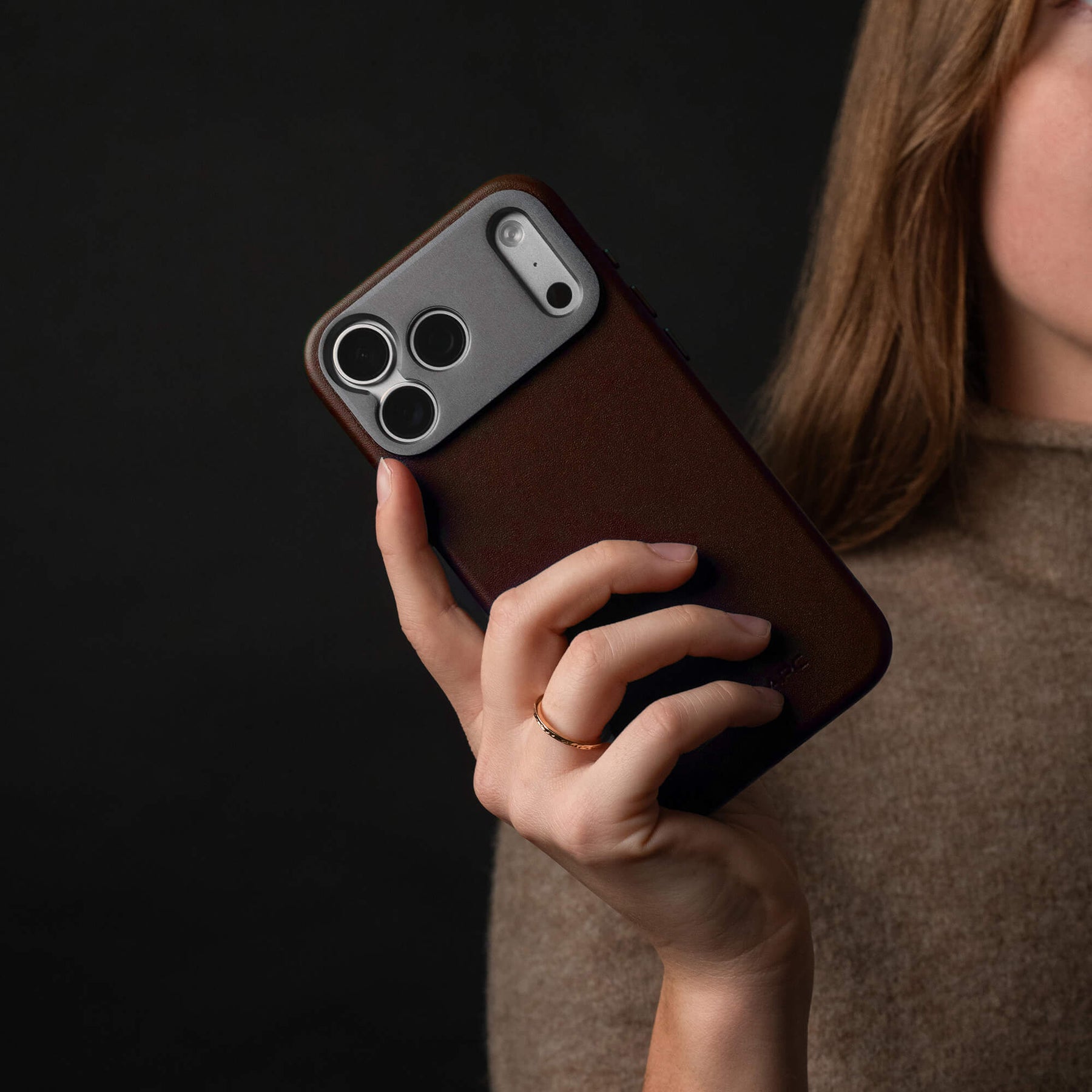 Woman holding an iPhone in the SANDMARC Pro Leather Case in brown against a dark background, wearing a gold ring and a taupe knit top, showing the leather texture and camera mount