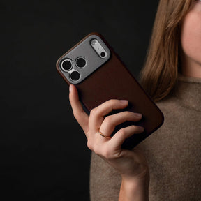 Woman holding an iPhone in the SANDMARC Pro Leather Case in brown against a dark background, wearing a gold ring and a taupe knit top, showing the leather texture and camera mount