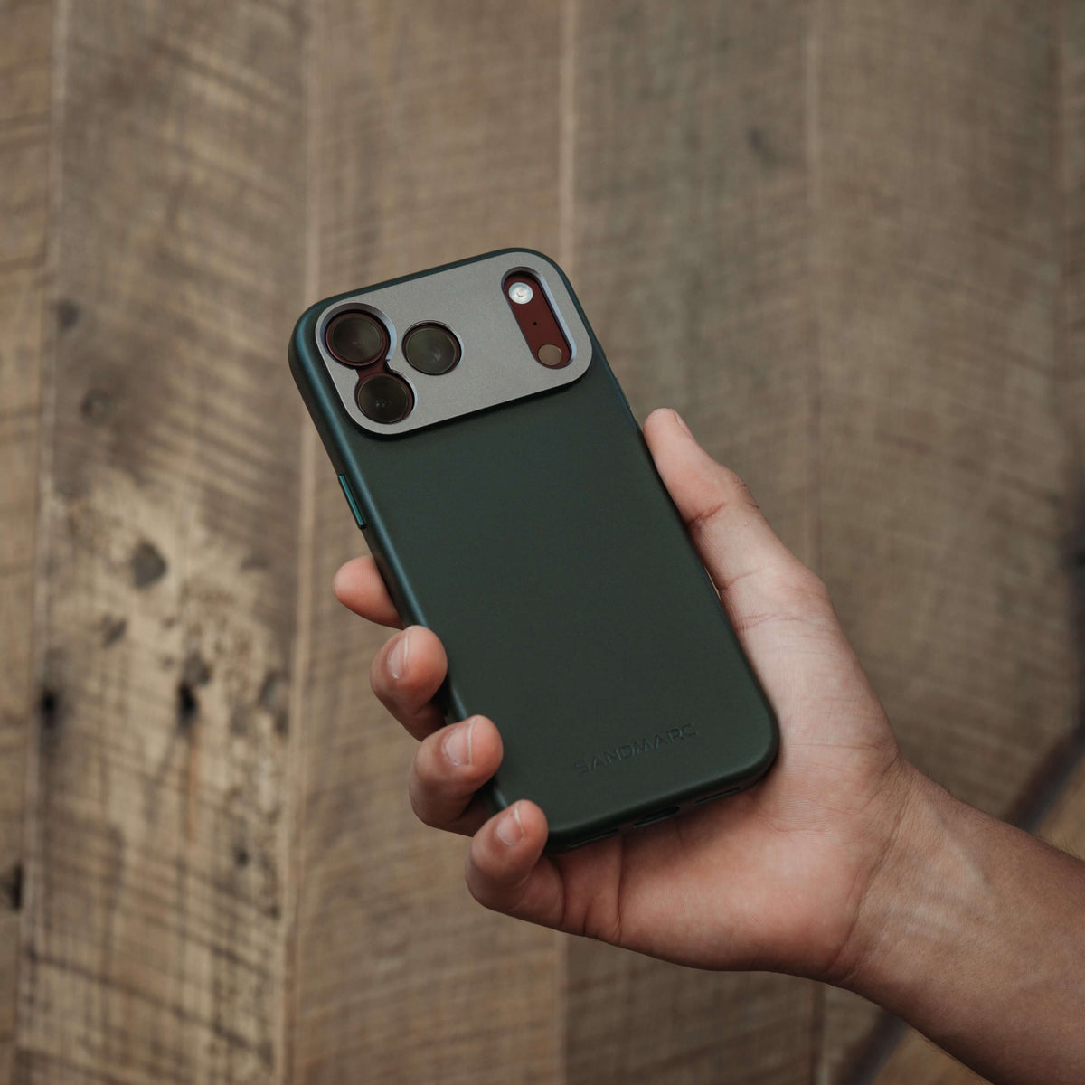SANDMARC Pro Leather Case for iPhone 18 Pro in Forest Green, held in a hand against a weathered wooden plank wall