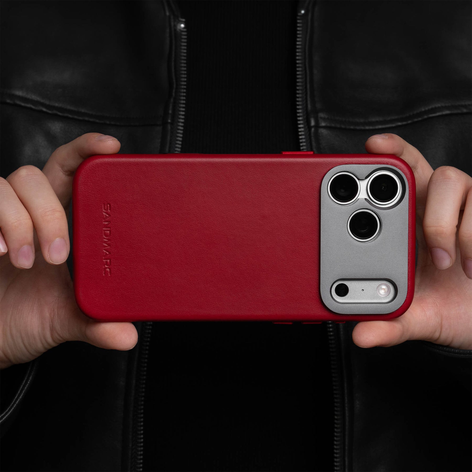 Two hands holding an iPhone in the SANDMARC Pro Leather Case in red horizontally in front of a black leather jacket, showing the SANDMARC logo embossed on the leather and triple camera cutout