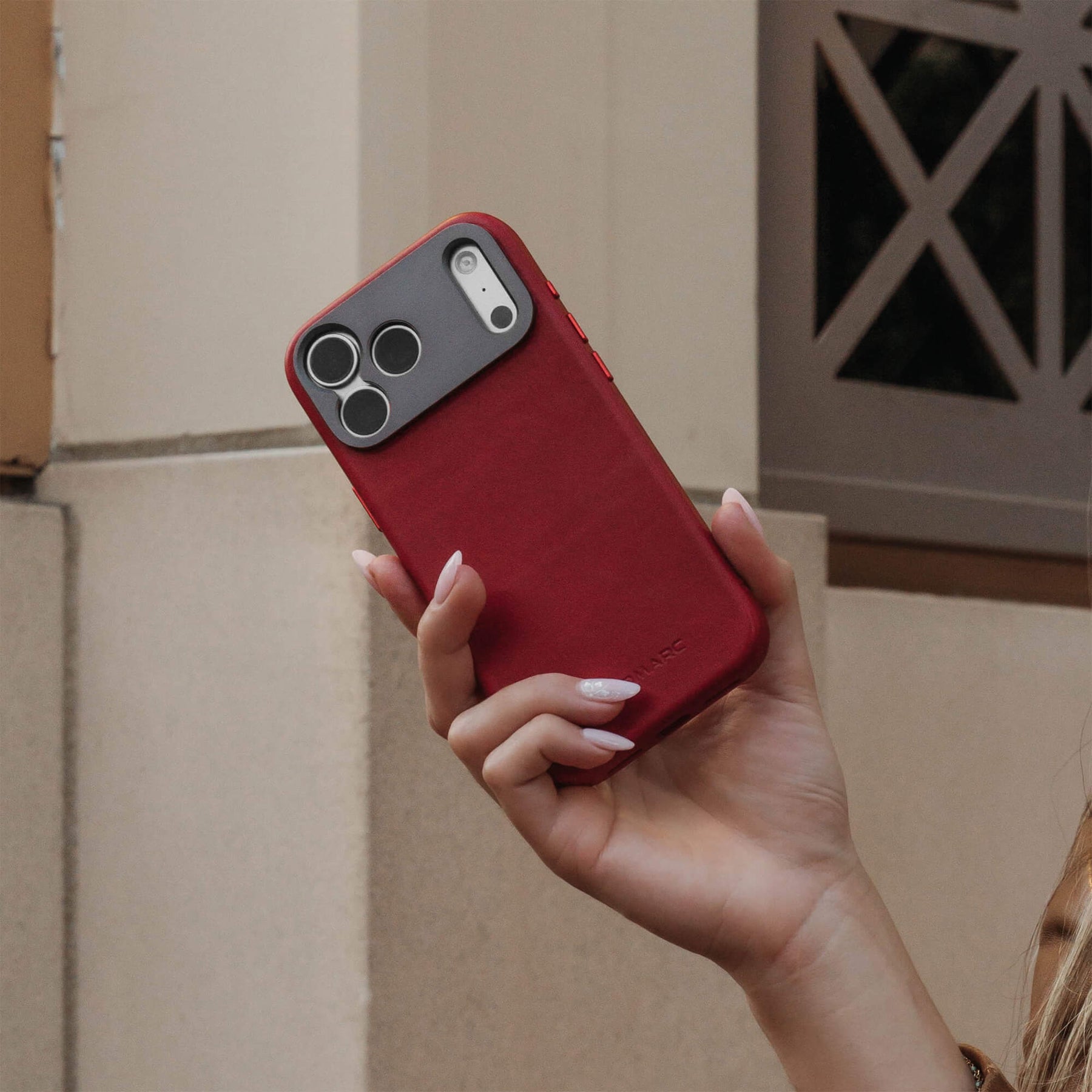 Woman's hand holding an iPhone in the SANDMARC Pro Leather Case in red outdoors against a beige stone building, showing the aluminum camera mount detail