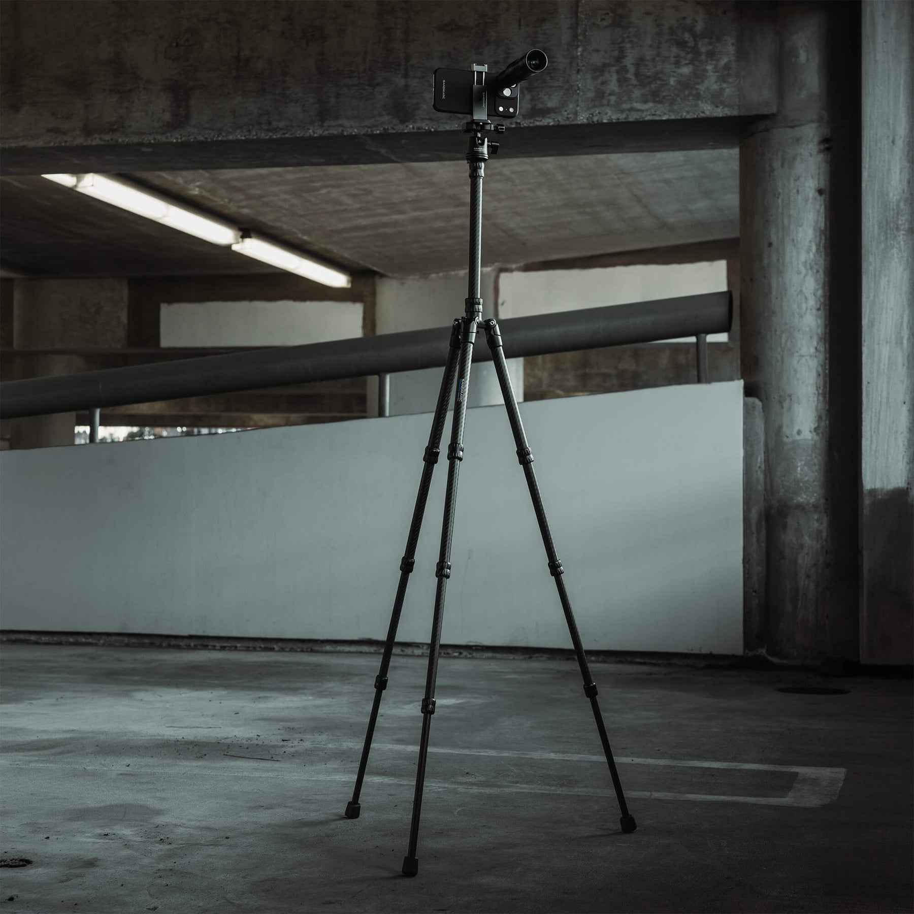 SANDMARC Carbon Fiber Tripod fully extended inside a brutalist concrete parking structure, with an iPhone 18 Pro Max mounted at the top fitted with the Telephoto 10x Lens, fluorescent lighting and concrete pillars visible in the background